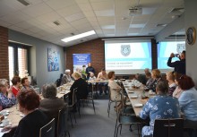 Seniorze poczuj się bezpiecznie. - Warsztaty o bezpieczeństwie dla seniorów.  Wykład policjantów. sala konferencyjna, seniorzy siedzą przy stołach, policjanci opowiadają, treść wyświetlona na ekranie. - powiększ