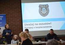 Seniorze poczuj się bezpiecznie. - Warsztaty o bezpieczeństwie dla seniorów.  Wykład policjantów. sala konferencyjna, seniorzy siedzą przy stołach, policjanci opowiadają, treść wyświetlona na ekranie. - powiększ