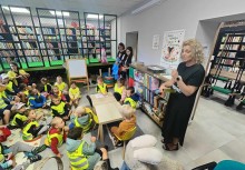 Wakacje z Urzędem Gminy Żukowo - Dzieci z Przedszkola z Miszewa w kamizelkach z opiekunkami, Biblioteka Samorządowa w Żukowie, regały z książkami, Pani Dyrektor Biblioteki Natalia Langa opowiada dzieciom o bibliotece. - powiększ