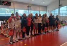 Grupa trenerów i organizatorów stoi wspólnie na hali sportowej podczas ceremonii zakończenia turnieju. W tle widoczni zawodnicy i kibice. Wręczane są podziękowania.  - powiększ