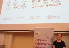 Na zdjęciu dziewczynka stojąca przy rollup-ie z napisem 'Olimpiada Matematyczna Juniorów' - powiększ