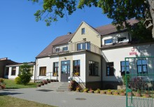 Budynek szkoły z jasną elewacją, ozdobiony kwiatami w donicach, sfotografowany w słoneczny dzień. - powiększ