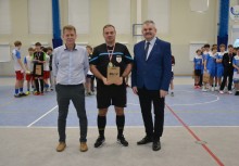 Trzech mężczyzn po meczu futsalu na hali widowiskowo-sportowej. Jeden z nich stojący na środku ma medal i trzyma prezent - powiększ