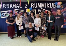 9 osób stojących przed 'ścianką' dekoracyjną z napisem 'Karnawał z Rummikub' - powiększ