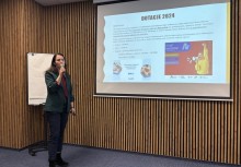 Na zdjęciu kobieta mówiąca przez mikrofon oraz przedstawiająca prezentację podczas spotkania - powiększ