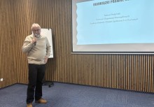 Na zdjęciu mężczyzna mówiący przez mikrofon oraz przedstawiający prezentację podczas spotkania - powiększ