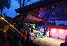 Grupa mieszkańców, w tym wiele dzieci, zgromadzona pod drewnianą altaną oświetloną kolorowymi reflektorami – wieczorny pokaz sztukmistrza Pablo podczas Nocy Muzeów w Gminie Żukowo. - powiększ