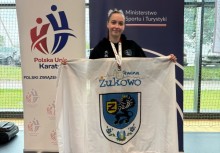 Zawodniczka z medalem pozuje z flagą Gminy Żukowo na tle roll-upów Polskiej Unii Karate i Ministerstwa Sportu i Turystyki. Zdjęcie wykonano w hali sportowej.