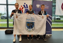 Troje młodych sportowców z medalami pozuje z flagą Gminy Żukowo na tle banerów Polskiej Unii Karate i Ministerstwa Sportu i Turystyki. Zdjęcie wykonano w hali sportowej po zawodach. - powiększ
