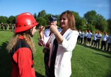 Kobieta w białej marynarce zakłada medal młodej druhnie w czerwonym hełmie podczas ceremonii na świeżym powietrzu. W tle widoczna jest szeregowa formacja uczestników wydarzenia oraz zielone otoczenie. - powiększ