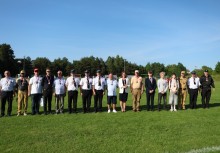 Grupa uczestników wydarzenia sportowo-strażackiego pozuje do wspólnego zdjęcia na boisku. Osoby w różnorodnych strojach – od mundurów strażackich po ubrania codzienne – stoją w szeregu na tle zieleni i błękitnego nieba. - powiększ