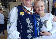 Starsza kobieta w tradycyjnym kaszubskim stroju ludowym pozuje z uśmiechniętą dziewczynką w białej bluzce. Obie stoją blisko siebie i obejmują się serdecznie. W tle widoczni są inni uczestnicy wydarzenia. - powiększ
