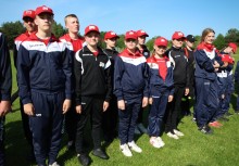 Grupa młodzieży w jednolitych sportowych strojach i czerwonych czapkach pozuje na zielonym boisku podczas zawodów lub uroczystości. Wszyscy stoją w szeregu, uśmiechnięci, na tle słonecznego nieba. - powiększ