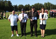 Cztery osoby pozuje do zdjęcia na boisku – dwóch strażaków z medalami i plakietami, towarzyszy im kobieta w białej marynarce oraz mężczyzna w jasnej koszuli. W tle widoczne są formacje uczestników wydarzenia i zielone otoczenie. - powiększ