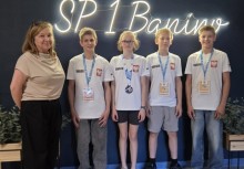 Czterech uczniów z medalami na szyi w towarzystwie dyrektor szkoły na tle ściany z neonowym napisem 'SP 1 Banino'. - powiększ