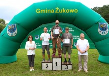 Trzech mężczyzn na podium, poza podium z lewej strony stoi kobieta a z prawej mężczyzna . - powiększ