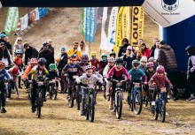 Grupa dzieci w kaskach jedzie na rowerach. W tle widać kibiców oraz banery promocyjne. 