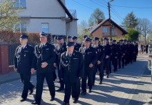 Strażacy w galowych mundurach podczas przemarszu. W tle widać innych uczestników wydarzenia oraz budynki mieszkalne.  - powiększ