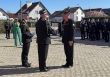 Dwaj strażacy w galowych mundurach podają sobie ręce. Jeden z nich ma na szyi medal na wstędze. Po lewej stronie stoi kolejny funkcjonariusz. W tle widać innych uczestników uroczystości.    - powiększ