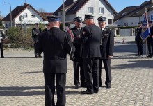 Na zdjęciu czterech strażaków, jeden z nich właśnie otrzymuje odznaczenie. W tle widać innych uczestników wydarzenia oraz budynki mieszkalne.  - powiększ