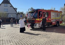 Ksiądz święci nowy wóz strażacki. Obok pojazdu stoi strażak w mundurze galowym. W te widać uczestników wydarzenia oraz budynki. - powiększ