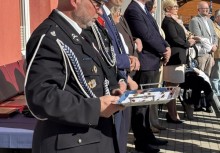 Strażak w mundurze galowym trzyma tacę z odznaczeniami. W tle widać szereg osób w strojach wizytowych.  - powiększ