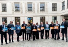 Grupa trzynastu osób stojących w rzędzie przed białym budynkiem z napisem 'SEJM RZECZPOSPOLITEJ POLSKIEJ'. Uczestnicy trzymają w dłoniach małe, prostokątne tabliczki w kolorach niebieskim i pomarańczowym z białymi napisami.  - powiększ