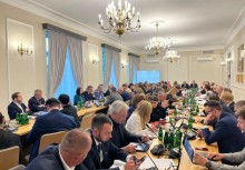 Sala konferencyjna wypełniona uczestnikami siedzącymi przy dwóch długich, drewnianych stołach. - powiększ
