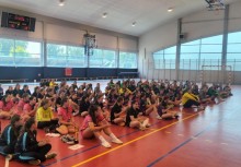 Zawodniczki z różnych drużyn siedzą na boisku w hali sportowej, ubrane w stroje klubowe. Trwa uroczystość zakończenia turnieju – atmosfera skupienia i oczekiwania. - powiększ
