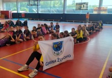 Zawodniczki siedzą w rzędach na hali sportowej podczas ceremonii zakończenia turnieju. Na pierwszym planie dziewczynka trzyma flagę z herbem i napisem „Gmina Żukowo”.  - powiększ