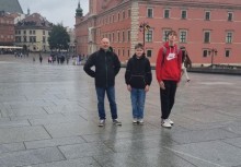 Trzech chłopców na tle Zamku Królewskiego w Warszawie – zdjęcie wykonane na placu Zamkowym w deszczowy dzień. Widoczna czerwona elewacja i wieże charakterystyczne dla warszawskiej Starówki. - powiększ