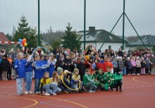 Kolorowe drużyny gotowe do sportowej rywalizacji – na nowo otwartym boisku w Leźnie grupy uczniów ubrane w barwne stroje (niebieskie, żółte, zielone i czerwone) pozują z obręczami olimpijskimi. W tle licznie zgromadzeni uczestników. - powiększ