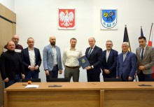 Na zdjęciu 9 osób w sali Rady Miejskiej w Urzędzie Gminy w Żukowie. Burmistrz trzyma teczkę.