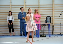 Uczennica występuje z mikrofonem podczas szkolnej uroczystości – w tle nauczyciele i dekoracja sali gimnastycznej. - powiększ