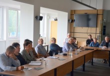 Zdjęcie przedstawia uczestników spotkania siedzących przy stole w jasnej sali konferencyjnej. Widoczne są notatniki, szklanki z wodą i ekran na ścianie. Atmosfera skupiona i robocza. - powiększ