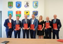7 osób, wszyscy trzymają teczki z herbem Gminy Żukowo. W tle na ścianie herby wszystkich gmin powiatu kartuskiego.  - powiększ
