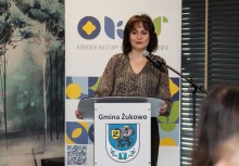 Na zdjęciu widoczna jest Burmistrz Gminy Żukowo, która stoi przy mównicy z herbem gminy i przemawia do mikrofonu. W tle znajduje się baner Ośrodka Kultury i Sportu.  - powiększ