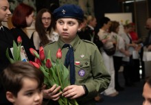 Na zdjęciu widać chłopaka w stroju harcerskim trzymającego w dłoniach tulipany. W tle widać innych uczestników wydarzenia.  - powiększ