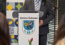 Na zdjęciu widoczny jest Radny Rady Miejskiej w Żukowie, który stoi przy mównicy z herbem gminy i przemawia do mikrofonu.  W tle znajduje się baner Ośrodka Kultury i Sportu.  - powiększ