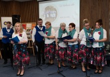 Na zdjęciu grupa śpiewających osób. W tle widoczna jest prezentacja multimedialna. - powiększ