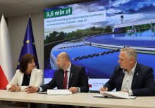 Kobieta i dwóch mężczyzn siedzą przy stole i podpisują dokumenty. W tle widać duży ekran z napisem '3,6 mln zł - Uporządkowanie gospodarki wodno-ściekowej na terenie Gminy Żukowo - etap III', flagę Polski oraz Unii Europejskiej. - powiększ