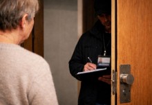 Na zdjęciu uchylone drzwi w których widać postać z notatnikiem i identyfikatorem 