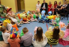 Dzieci w kolorowych przebraniach siedzą w kręgu. W tle widać dorosłych siedzących na krzesłach.  - powiększ