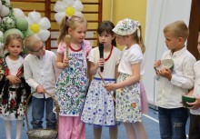 Na pierwszym planie trzy dziewczynki w fartuszkach z motywem kaszubskim. Jedna z dziewczynek mówi do mikrofonu. Obok nich stoją dzieci.  - powiększ