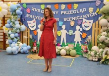 Kobieta przemawia do mikrofonu. W tle widać ściankę dekoracyjną z balonami, napisem 'XVII Gminny Przegląd Małych Form Scenicznych',  maskami teatralnymi oraz sylwetkami dzieci.  - powiększ