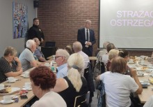 Spotkanie dla seniorów, starsi ludzie za stołem, ekran z wyświetlonymi informacjami, Burmistrz Gminy Żukowo Wojciech Kankowski. - powiększ