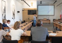 Uczestnicy spotkania siedzą przy stole na Sali Rady Miejskiej, jeden mężczyzna stoi. W tle widać ekran projekcyjny.  - powiększ