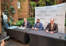 Trzy osoby przy stole podpisują umowę. Za nimi ścianka konferencyjna - powiększ