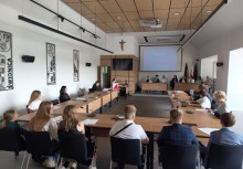 Młodzi ludzie siedzą przy stołach ustawionych w podkowę w dużej sali konferencyjnej