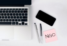 Laptop, dwa długopisy, smartfon oraz różowa karteczka z napisem: NGO